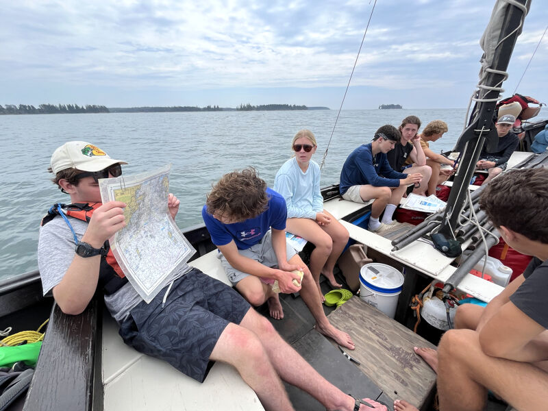 The photo shows a group of people on a sailboat. One person is holding a map and appears to be navigating. Others are sitting and relaxing. The sky is overcast, and the water is calm. It looks like a leisurely day on the water.